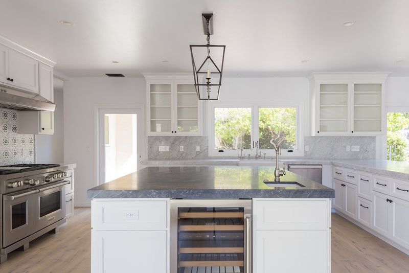 A bright, open custom kitchen featuring white shaker cabinets, a large center island with dark marble countertop and built-in wine cooler, stainless steel range, and glass-front upper cabinets flanking garden-view windows.