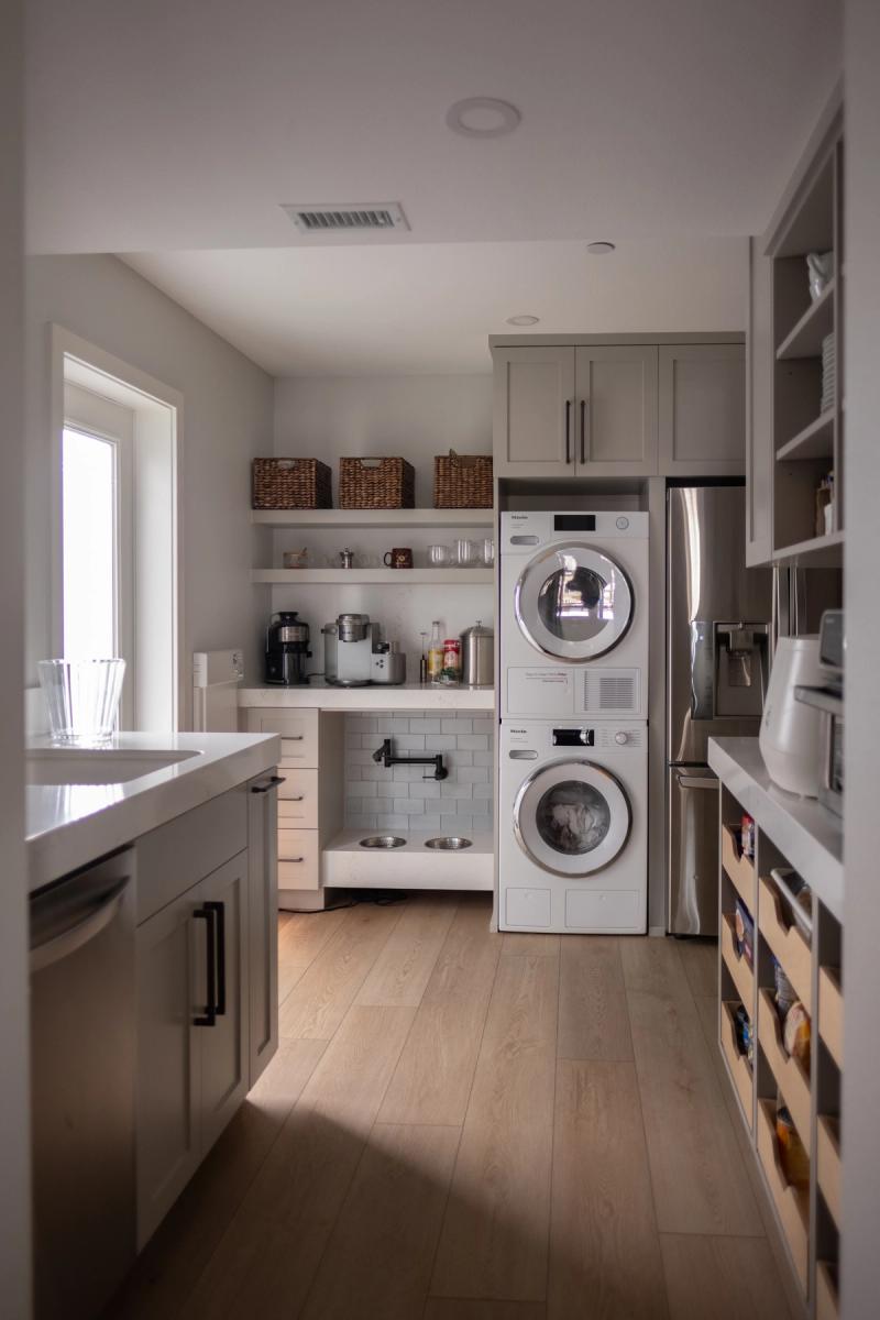 A well-appointed butler's pantry featuring gray shaker cabinetry, open shelving with wicker baskets, a built-in coffee bar station, stacked Miele washer and dryer, and a pet feeding station with a pot filler, all finished with light wood floors and white quartz countertops.