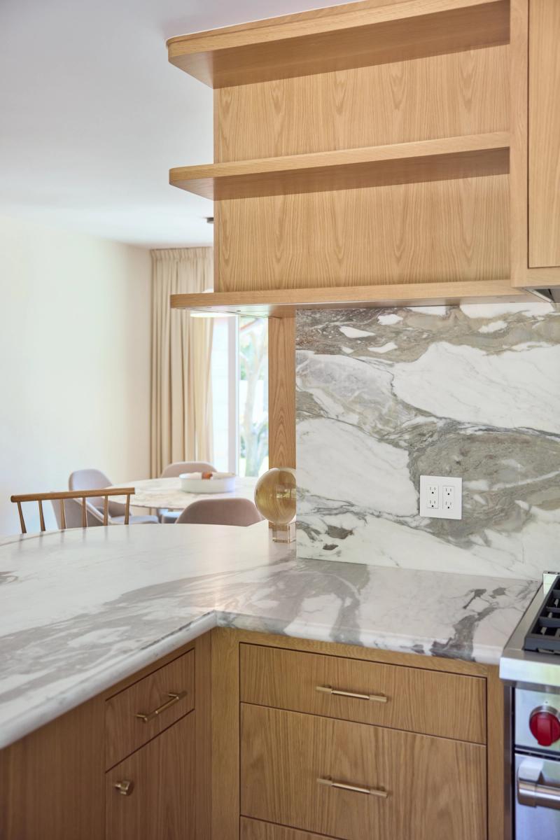 A modern kitchen featuring warm white oak cabinetry with brass hardware, dramatic marble countertops and backsplash, and floating open shelves. The space opens to a dining area in the background.