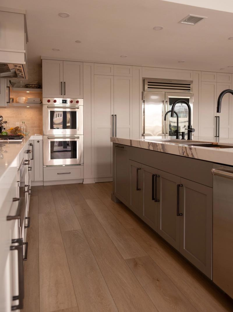 A spacious transitional kitchen featuring white upper cabinetry, a gray island with marble countertop, stainless steel double wall ovens, a built-in refrigerator, and wide-plank hardwood floors.