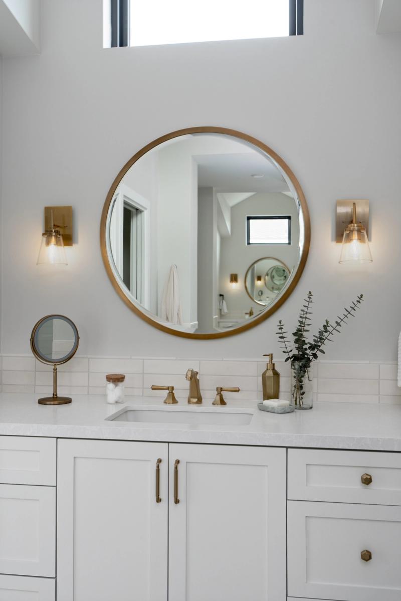 A elegant white shaker-style bathroom vanity with brushed brass hardware, a large round gold-framed mirror, flanking wall sconces, and a white quartz countertop with undermount sink.