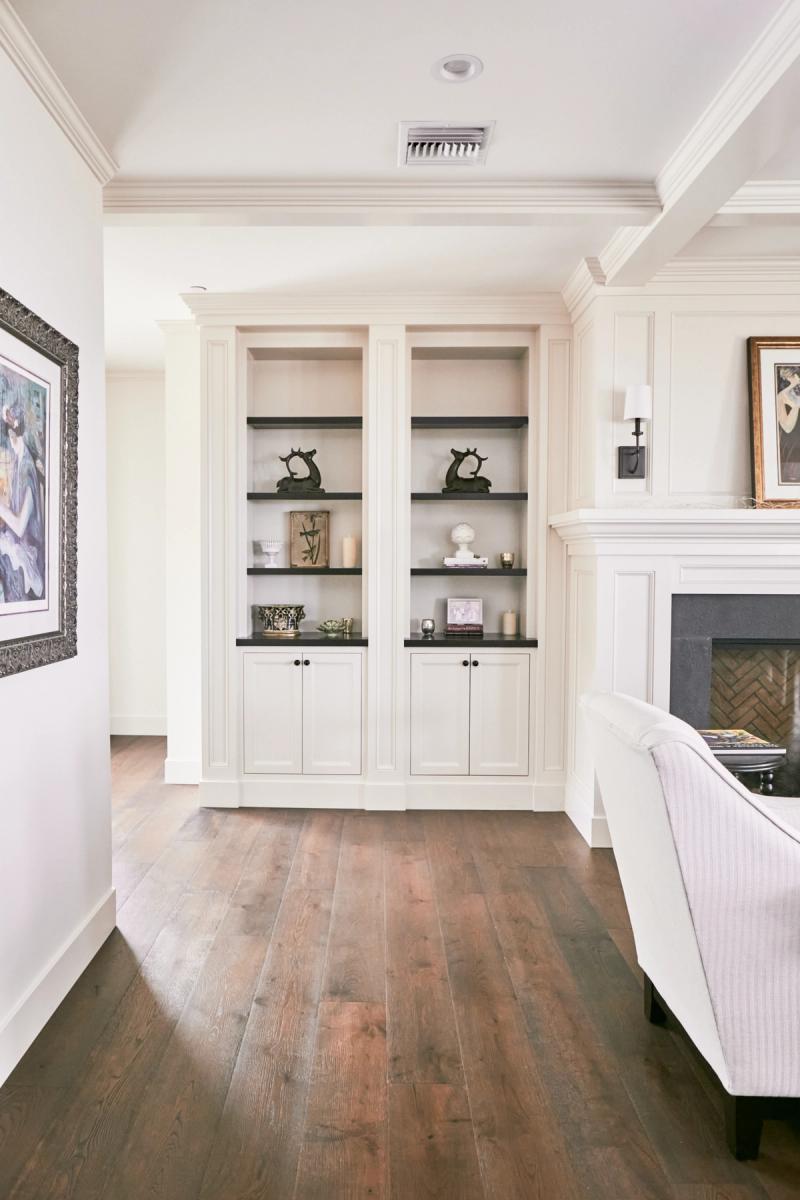 A pair of symmetrical white built-in bookcases with dark shelves and lower cabinet doors flank a hallway in a traditional living room, decorated with sculptural accents and candles, alongside a herringbone-tile fireplace.