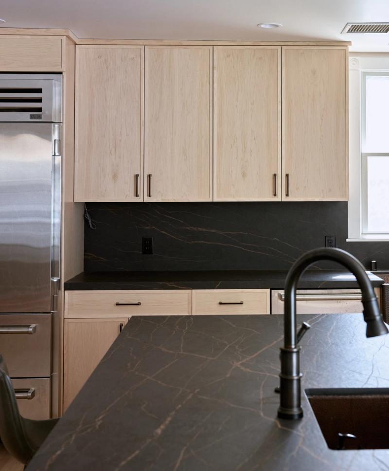 A modern kitchen featuring light natural oak cabinetry with matte black hardware, dramatic dark stone countertops and backsplash with copper veining, a matte black gooseneck faucet, and a stainless steel built-in refrigerator.