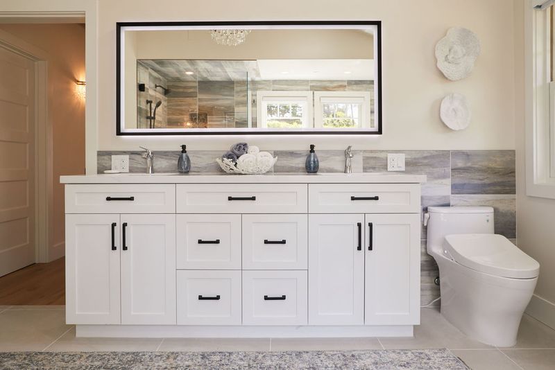 A wide double-sink bathroom vanity with white shaker-style cabinets, black hardware, marble backsplash, and a large framed mirror reflecting a walk-in shower. A modern toilet is visible to the right.
