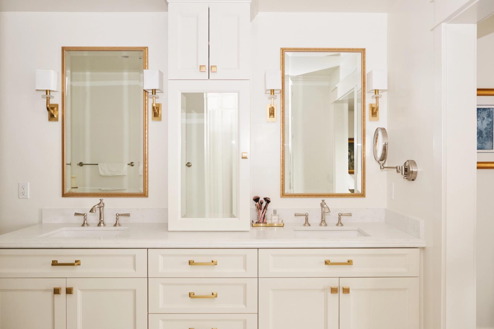 A bright, airy master bathroom featuring a white shaker-style vanity with brass hardware, a round gold-framed mirror, freestanding soaking tub, patterned mosaic floor tile, and vaulted ceiling with exposed beams.