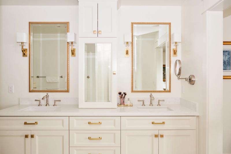 A bright, airy master bathroom featuring a white shaker-style vanity with brass hardware, a round gold-framed mirror, freestanding soaking tub, patterned mosaic floor tile, and vaulted ceiling with exposed beams.