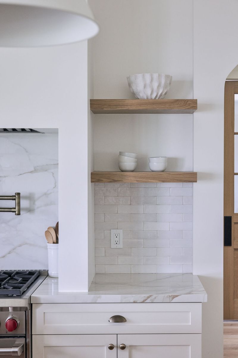 A bright, clean kitchen corner featuring two floating walnut shelves built into a niche, styled with white ceramic bowls, above a marble tile backsplash and white shaker cabinetry with a marble countertop.
