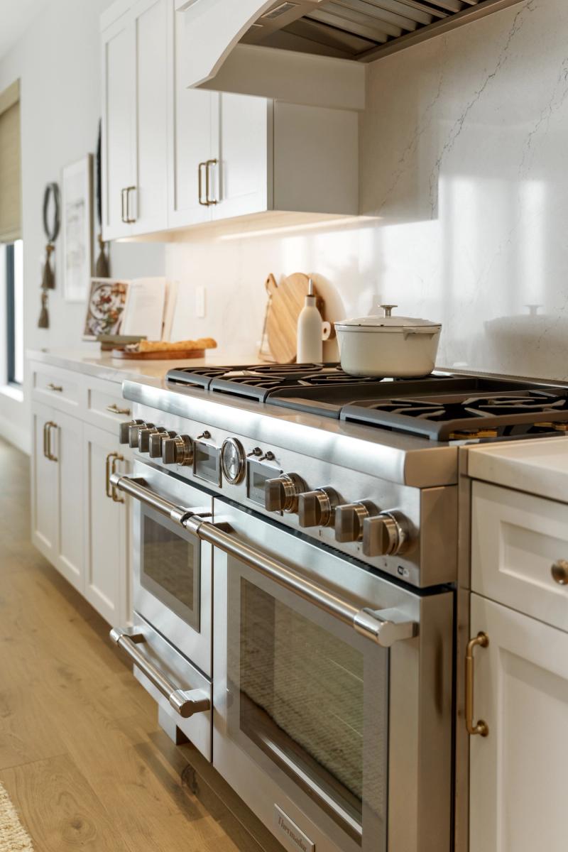 A bright, elegant kitchen featuring white shaker cabinets with brass hardware, a stainless steel professional dual-oven range with multiple burners, a marble slab backsplash, and a custom arched range hood. A white Dutch oven sits on the cooktop.