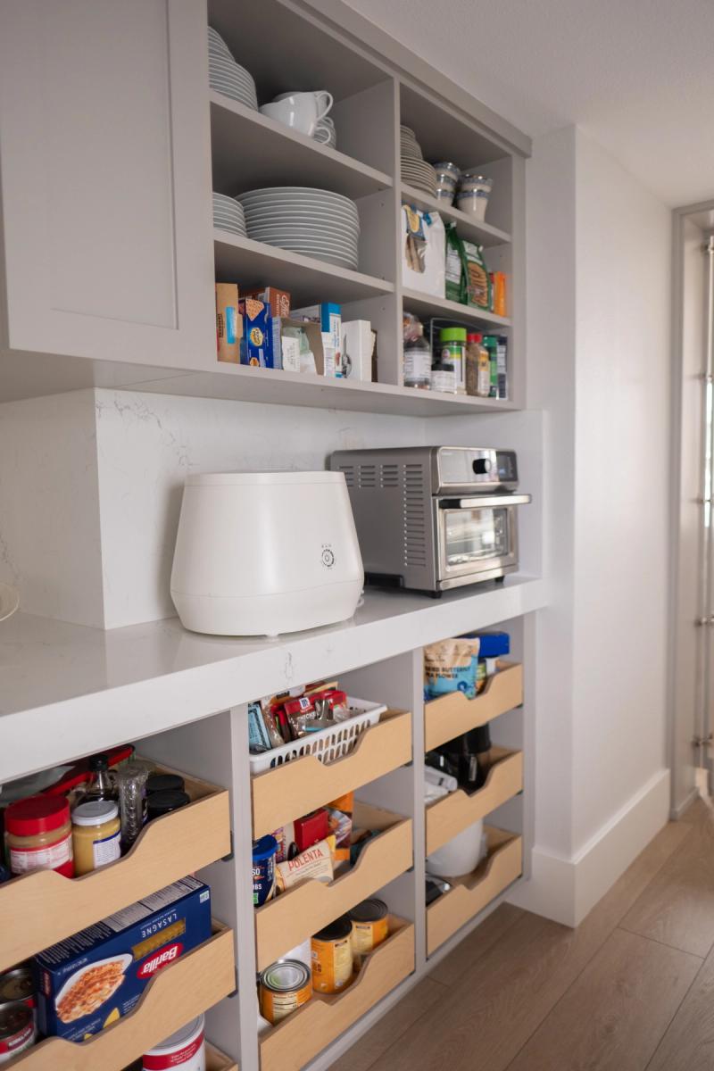 A well-organized custom pantry with gray painted cabinetry featuring open upper shelves holding dishes and pantry staples, a countertop with small appliances, and lower pull-out wooden drawers stocked with food items.