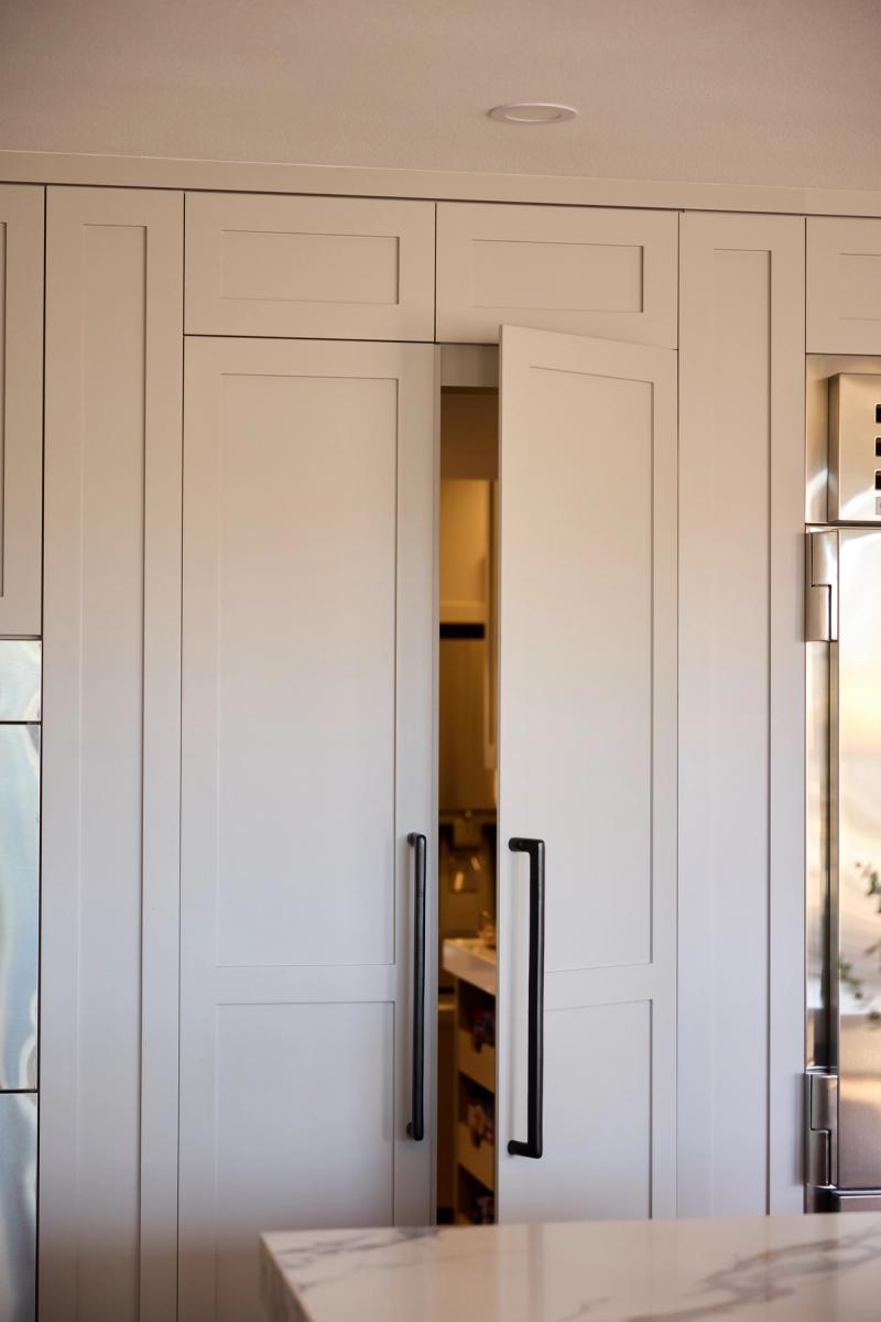 A wall of floor-to-ceiling painted shaker-style cabinetry in soft white with matte black pulls, one door slightly ajar revealing a hidden walk-in pantry behind it. A stainless steel refrigerator is partially visible to the right.