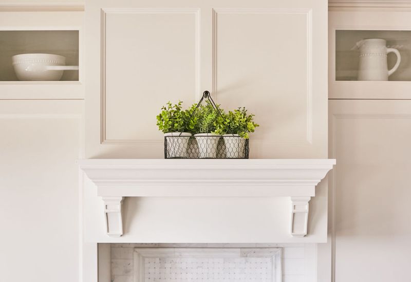 A bright white fireplace mantel with decorative corbels topped with a wire basket of green plants, flanked by glass-front built-in cabinetry displaying white dishware.