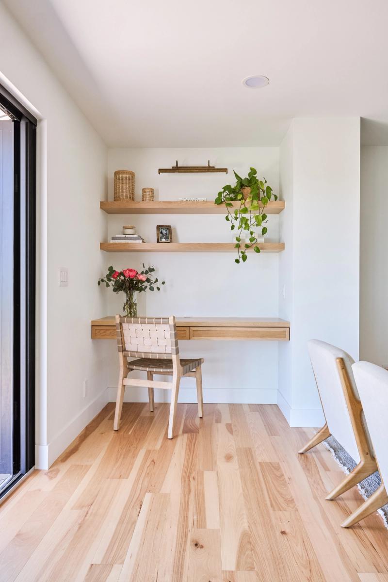 A minimalist home office nook featuring a floating white oak desk with drawers, two matching wall-mounted shelves styled with plants and woven baskets, and a woven leather chair on light hardwood floors.