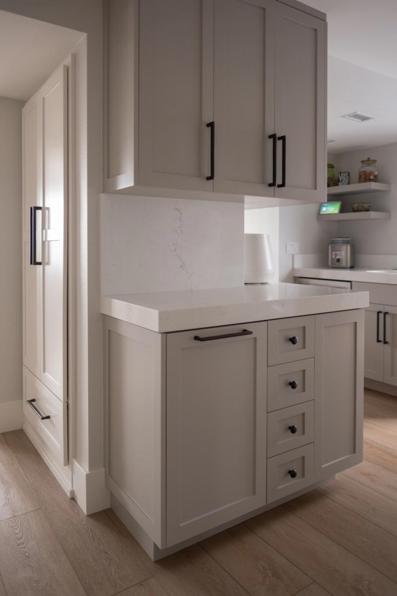 A transitional kitchen featuring greige shaker-style upper and lower cabinets with matte black hardware, white quartz countertops and backsplash, and light wood flooring. The layout includes a bank of drawers beside a lower cabinet door, with upper cabinets above and a pantry visible to the left.