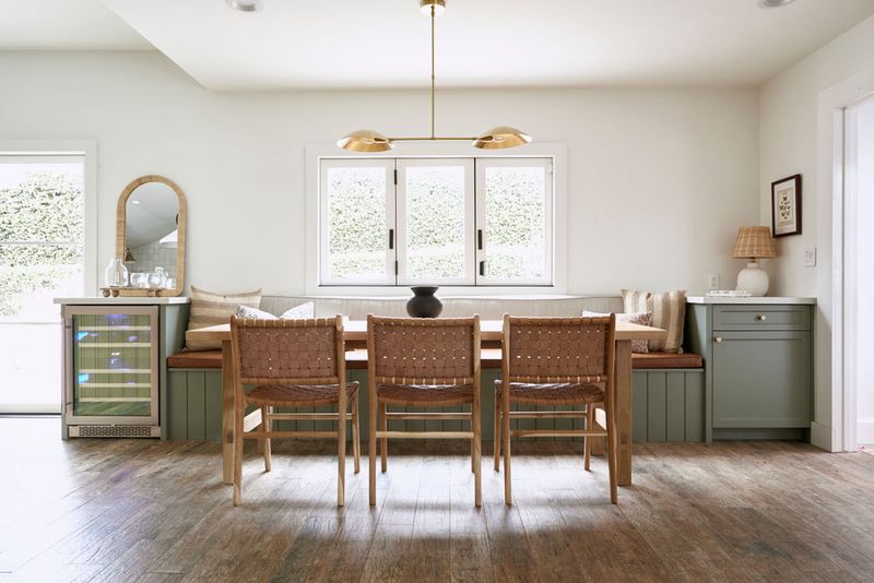A bright dining nook featuring custom sage green built-in bench seating with cushions, a wood dining table surrounded by woven leather chairs, and a built-in wine refrigerator, all beneath a gold three-light pendant chandelier.