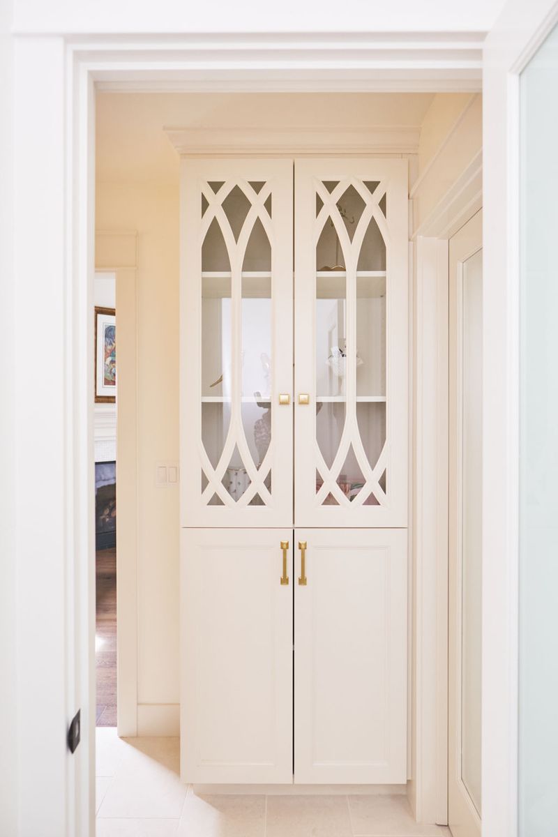 A tall cream-painted built-in cabinet with decorative Gothic arch glass upper doors and solid lower doors, accented with brushed brass hardware, set into a hallway nook.