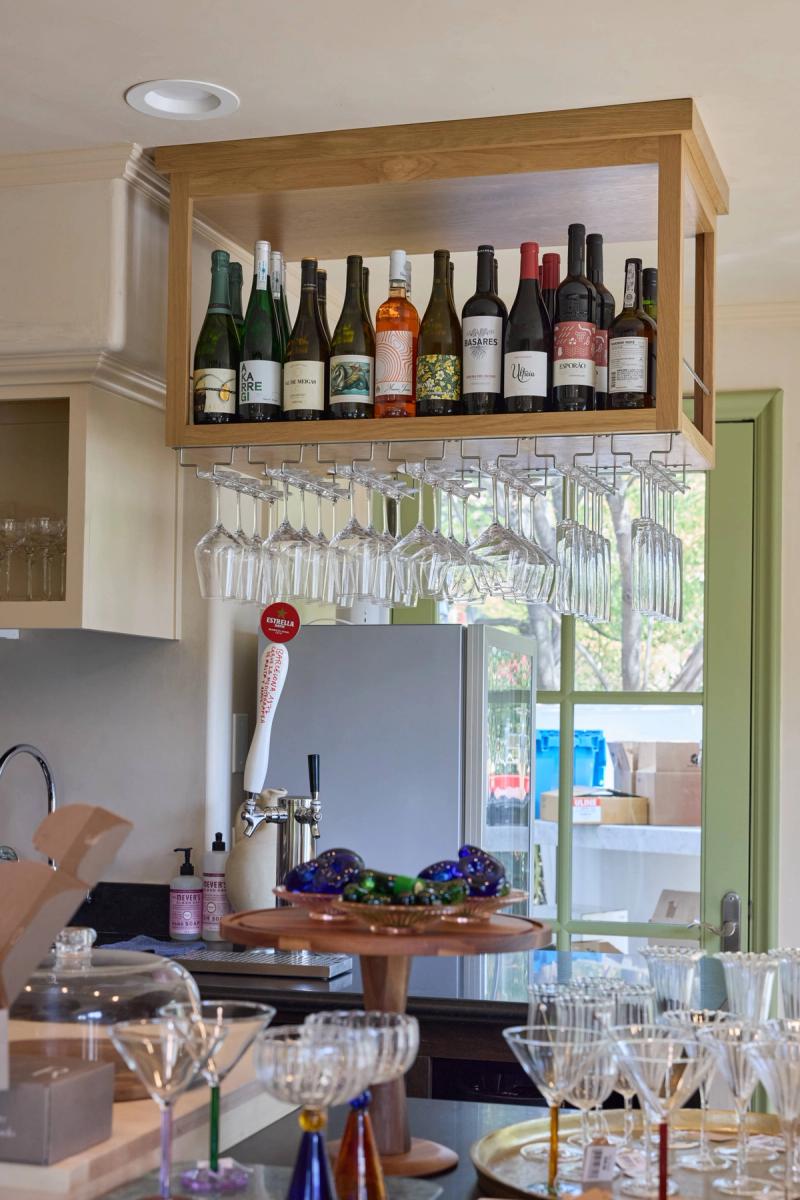 A custom oak overhead wine cabinet with built-in hanging stemware rack, displaying a collection of wine bottles above a bar area with a draft beer tap and assorted glassware.