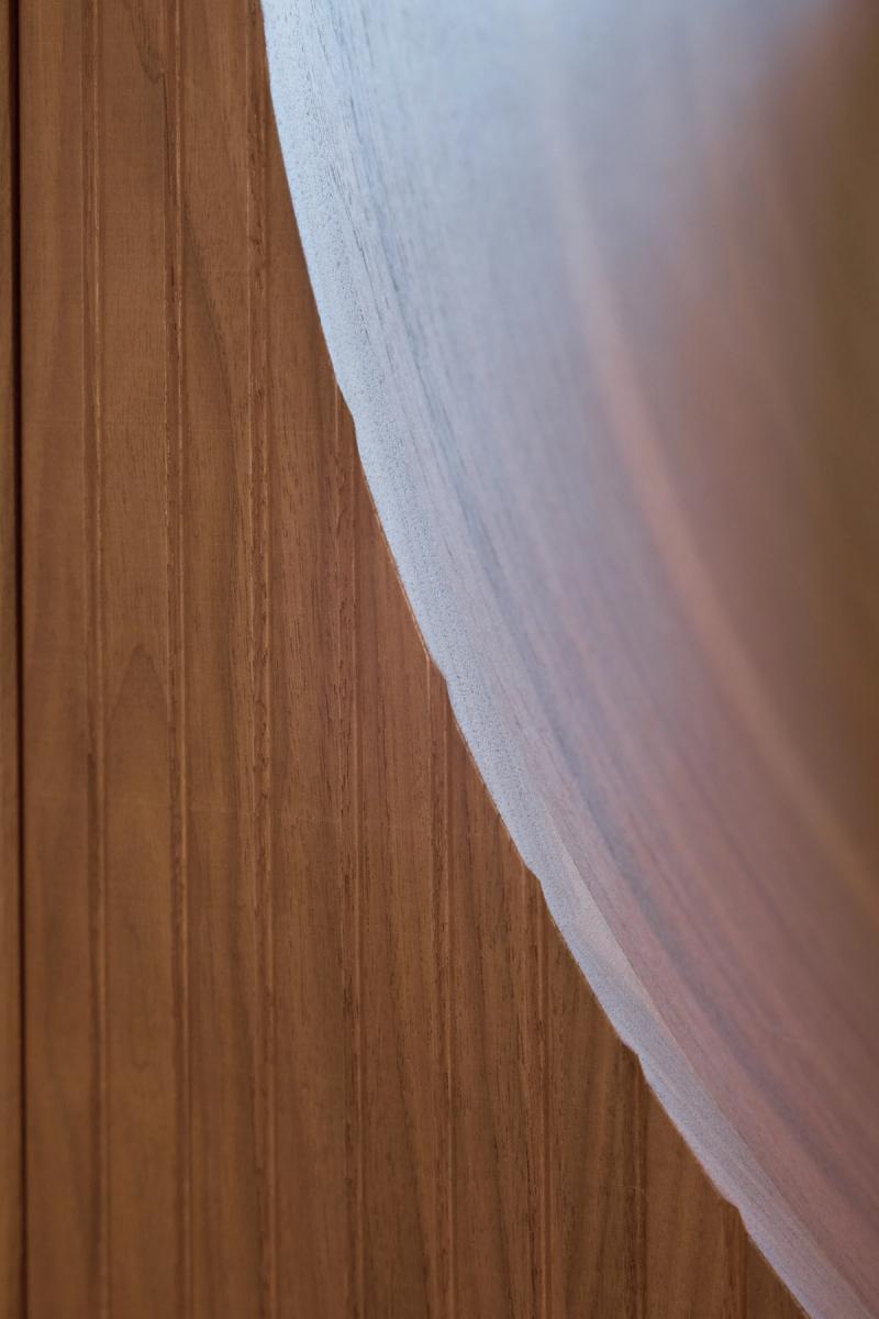 A close-up detail shot showing the curved edge of a frosted or etched glass panel meeting a warm wood grain cabinet surface. The image highlights the contrast between the smooth translucent glass and the rich natural wood texture.