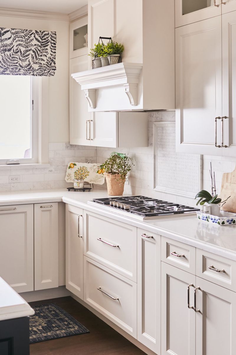 A bright, elegant kitchen featuring cream-painted inset cabinetry with brushed nickel hardware, white quartz countertops, a marble tile backsplash, and a decorative range hood above a gas cooktop.