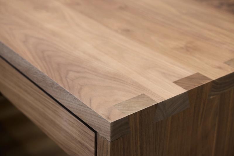 A close-up of the corner of a solid walnut wood surface showing fine grain detail, a clean mitered edge, and exposed finger joint construction on the leg.
