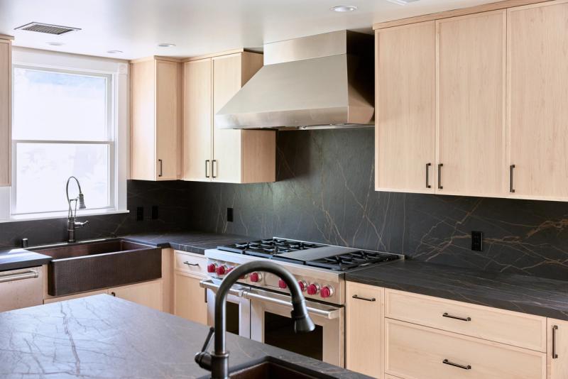 A modern kitchen featuring light maple flat-panel cabinetry, dramatic dark soapstone countertops and backsplash, a hammered copper farmhouse sink, and a professional stainless steel range with red knobs beneath a large vent hood.