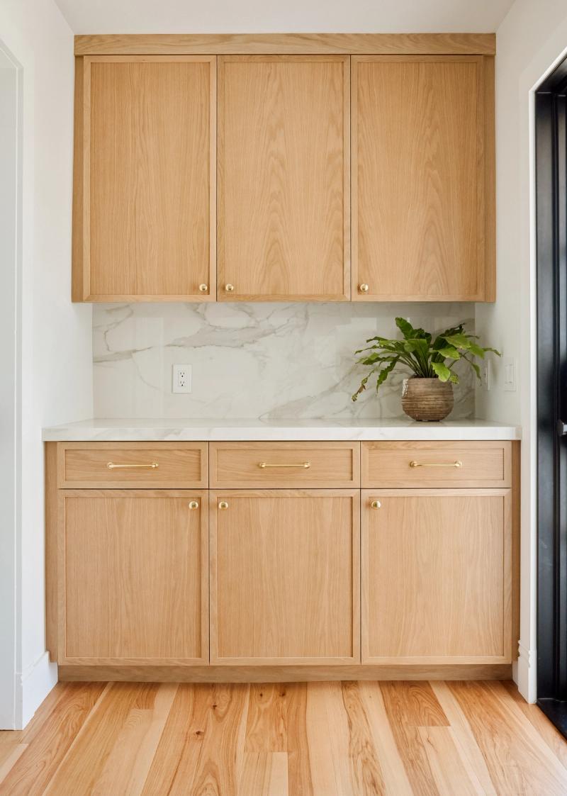 A butler's pantry featuring light white oak shaker-style cabinets with brass hardware, a white marble backsplash and countertop, and a potted green plant on the counter.