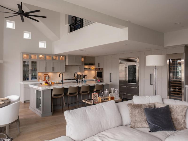A spacious open-concept kitchen featuring gray shaker cabinets, a large marble-top island with bar seating, stainless steel appliances including a built-in refrigerator and wine cooler, and a connected living area with vaulted ceilings.