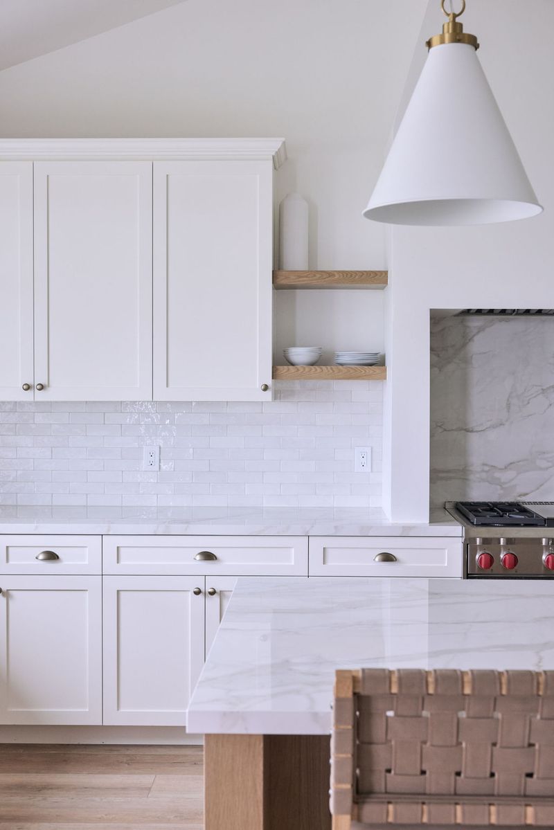 A bright, all-white custom kitchen featuring inset shaker cabinets, marble countertops, glossy subway tile backsplash, floating wood shelves, and a professional gas range with marble surround.