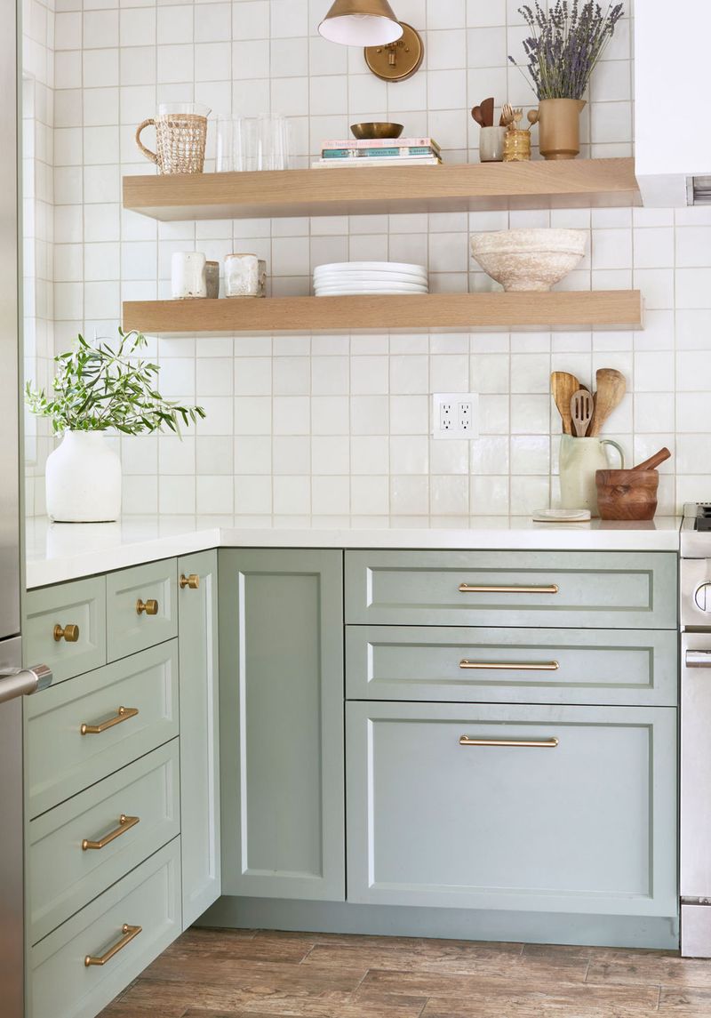 A bright kitchen featuring sage green shaker-style lower cabinets with brass hardware, white square tile backsplash, and natural wood floating shelves styled with ceramics and greenery.