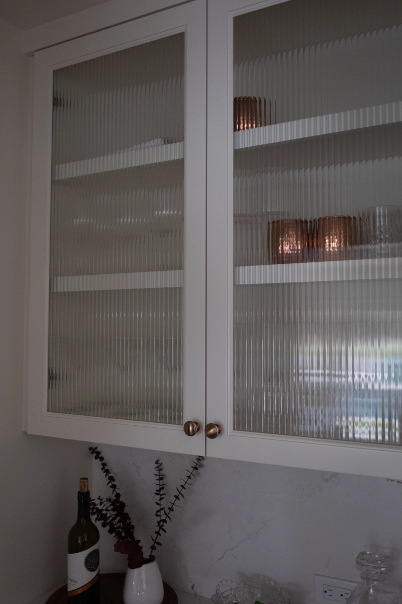 A pair of white upper kitchen cabinets with vertical reeded glass panel doors and brushed brass knobs, displaying pink glassware on interior shelves, with a marble backsplash and wine bottle visible below.