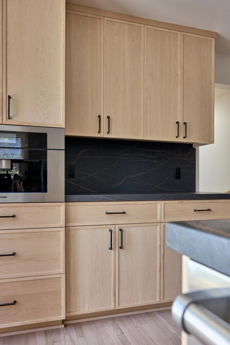 A modern kitchen featuring light maple flat-panel cabinets with matte black hardware, a dramatic dark marble-look stone backsplash, dark countertops, and a built-in Miele appliance.