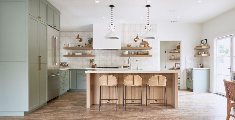A bright, open kitchen featuring sage green shaker cabinets, a natural wood island with three rattan barstools, white square tile backsplash, open wood shelving, and two white pendant lights.