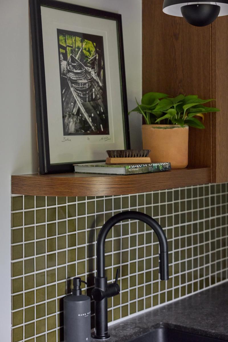 A close-up of a kitchen sink area featuring olive green mosaic tile backsplash, a matte black faucet and soap dispenser, a warm walnut floating shelf, and decorative styling with a framed print and potted plant.