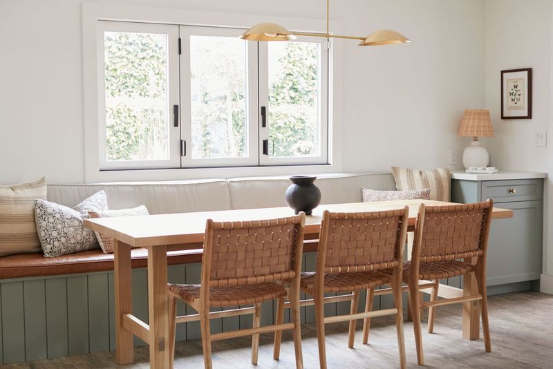 A bright dining nook featuring a custom sage green built-in banquette with leather cushions and decorative pillows, paired with a light wood dining table and woven leather chairs beneath a brass pendant light.