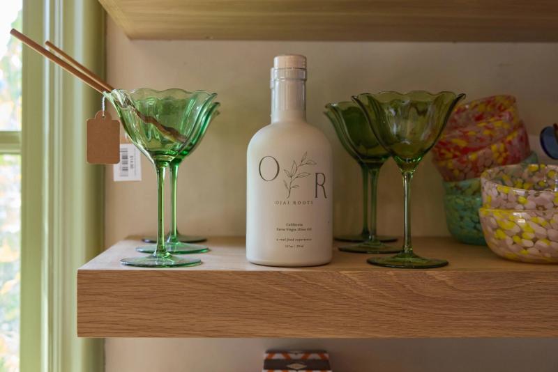 A close-up of a natural oak floating shelf displaying green glass coupe glasses, a bottle of Ojai Roots olive oil, and colorful glass bowls, set against a white wall with a green-trimmed window.