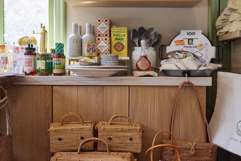 A warm oak wood counter and cabinet display in a specialty shop or kitchen showroom, featuring gourmet food products, olive oil bottles, a paella kit, and woven rattan baskets arranged beneath the counter.