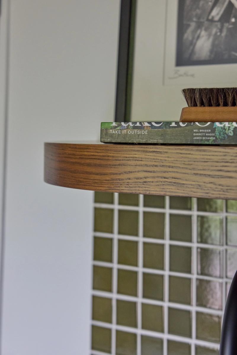 A close-up of a round walnut-stained wood side table top holding a green book titled 'Take It Outside' by Mel Brasier, Garrett Magee, and James DeSantis, with a wooden brush resting on top. A framed black and white photo and a grid-patterned cabinet or screen are visible in the background.