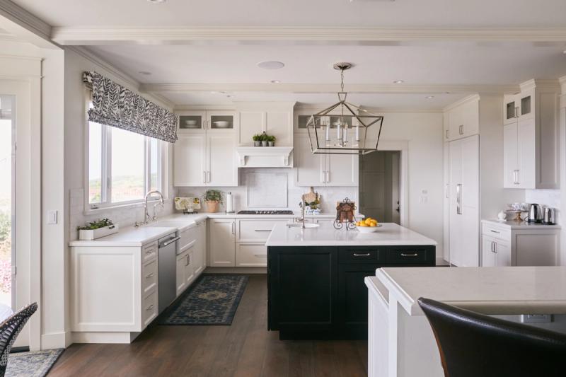 A bright, spacious traditional kitchen featuring white shaker-style cabinetry, a dark navy-black island with white quartz countertop, hardwood floors, and a geometric polished nickel pendant light above the island.