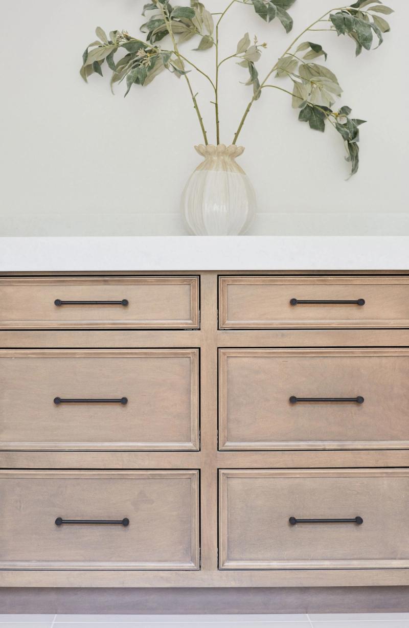 A close-up of a six-drawer bathroom vanity in light natural wood with raised panel fronts and matte black bar pulls, topped with a white stone countertop and styled with a ribbed glass vase holding eucalyptus branches.