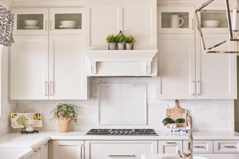 A bright, elegant white kitchen featuring custom inset cabinetry with glass uppers, a decorative wood range hood surround, marble tile backsplash, and quartz countertops accented with fresh greenery and silver hardware.
