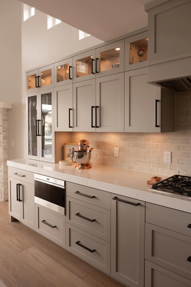 A modern transitional kitchen featuring warm gray shaker-style cabinetry with matte black hardware, white quartz countertops, a beige brick backsplash, illuminated glass upper cabinets, and a copper KitchenAid stand mixer on the counter.