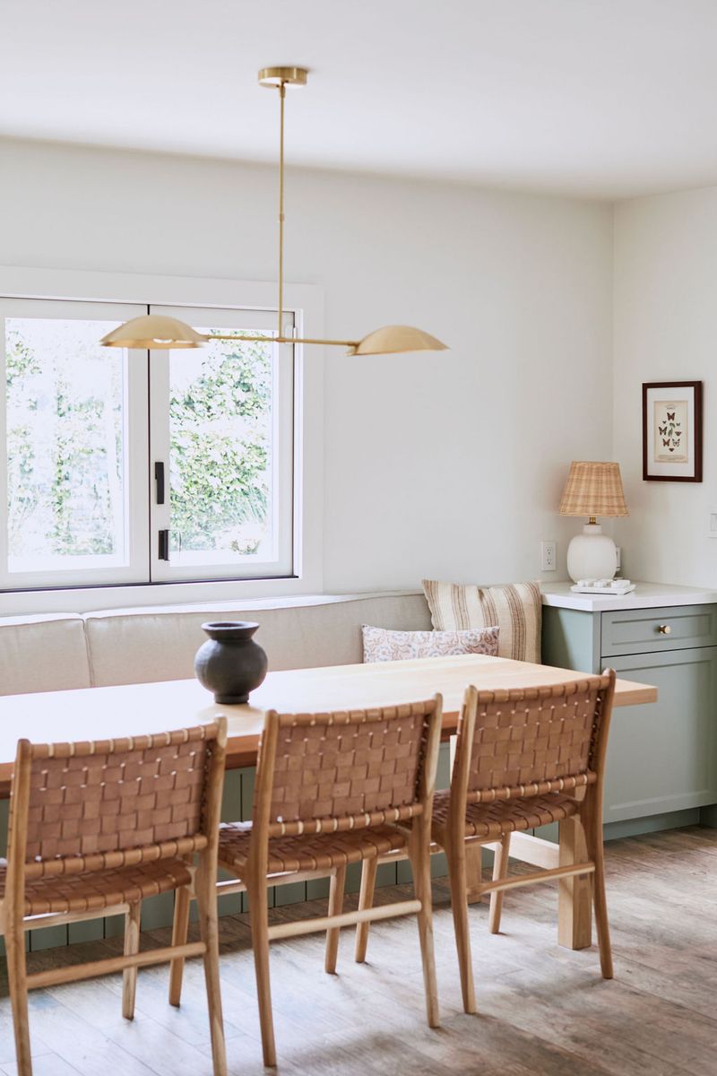A bright dining nook featuring sage green built-in bench seating with storage drawers, a light wood dining table, woven leather chairs, and a brass two-shade pendant light overhead.
