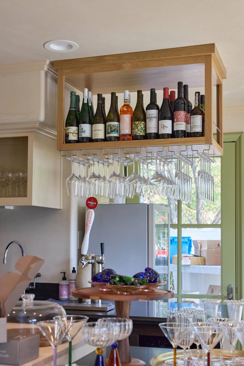 A custom wood overhead bar cabinet in natural oak displays a collection of wine bottles, with hanging wine glass racks below and a draft beer tap on the counter beneath.
