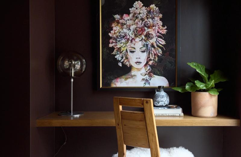A moody home office nook featuring a floating natural wood desk and matching wood chair against deep charcoal walls, styled with a globe table lamp, potted plant, stacked books, and a floral portrait painting in a gold frame.