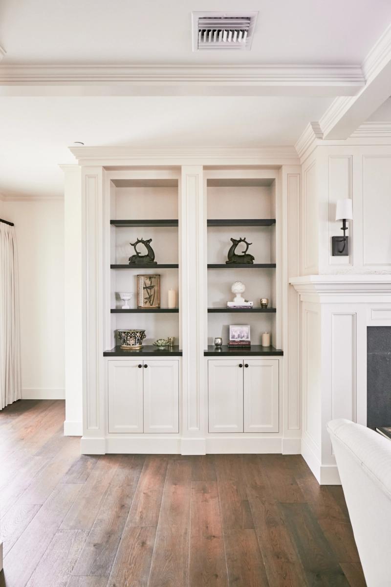 A pair of symmetrical cream-painted built-in bookcases with dark shelves and lower cabinet storage, styled with decorative objects, flanking a fireplace in a traditional living room with wide-plank hardwood floors.