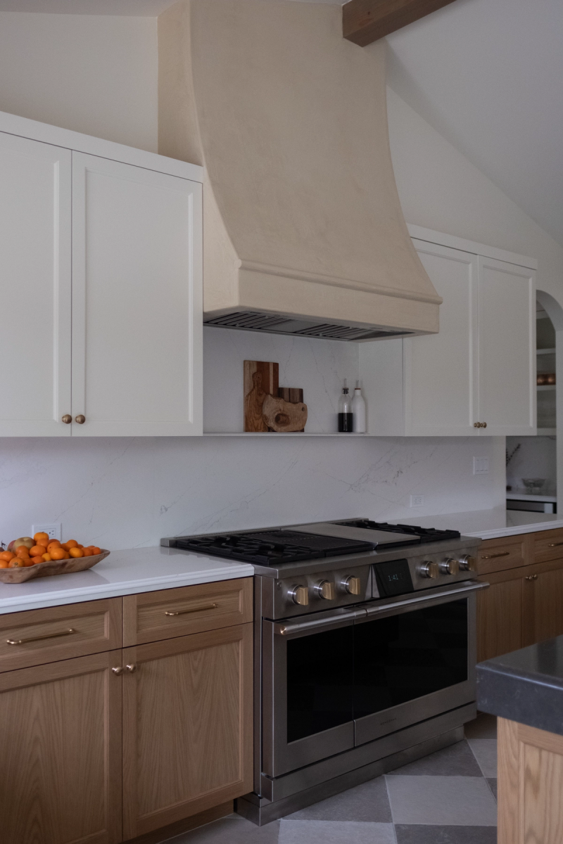 A custom kitchen featuring warm wood lower cabinets, white upper cabinets, a stainless steel range, marble backsplash, and a sculptural plaster range hood with an exposed wood ceiling beam above.