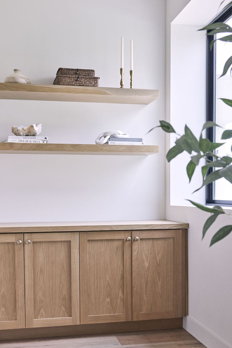 A light-filled built-in featuring white oak lower cabinets with a white countertop and two floating wood shelves styled with books, candles, woven baskets, and decorative objects beside a black-framed window.