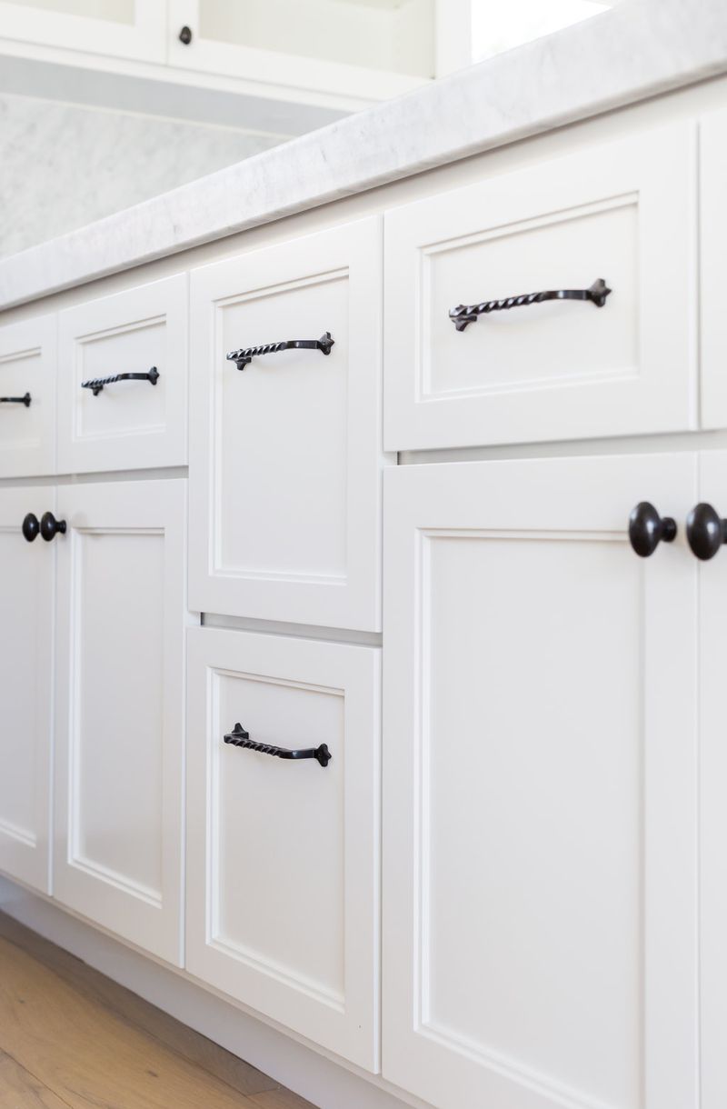 Close-up of white painted bathroom vanity cabinetry featuring raised panel doors and drawers with twisted black iron pull handles and round black cabinet knobs, topped with a white marble countertop.