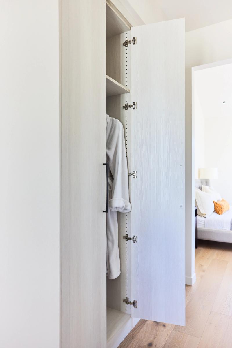 A partially open tall white wardrobe cabinet with chrome soft-close hinges and a matte black handle, with a white robe hanging inside and a bedroom visible in the background.