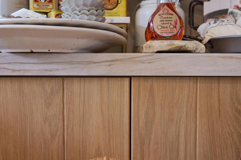 A close-up view of custom natural oak kitchen cabinets with clean flat-front drawer and door fronts, topped with a veined stone countertop and various kitchen items including a bottle of Calabrian chili olive oil.