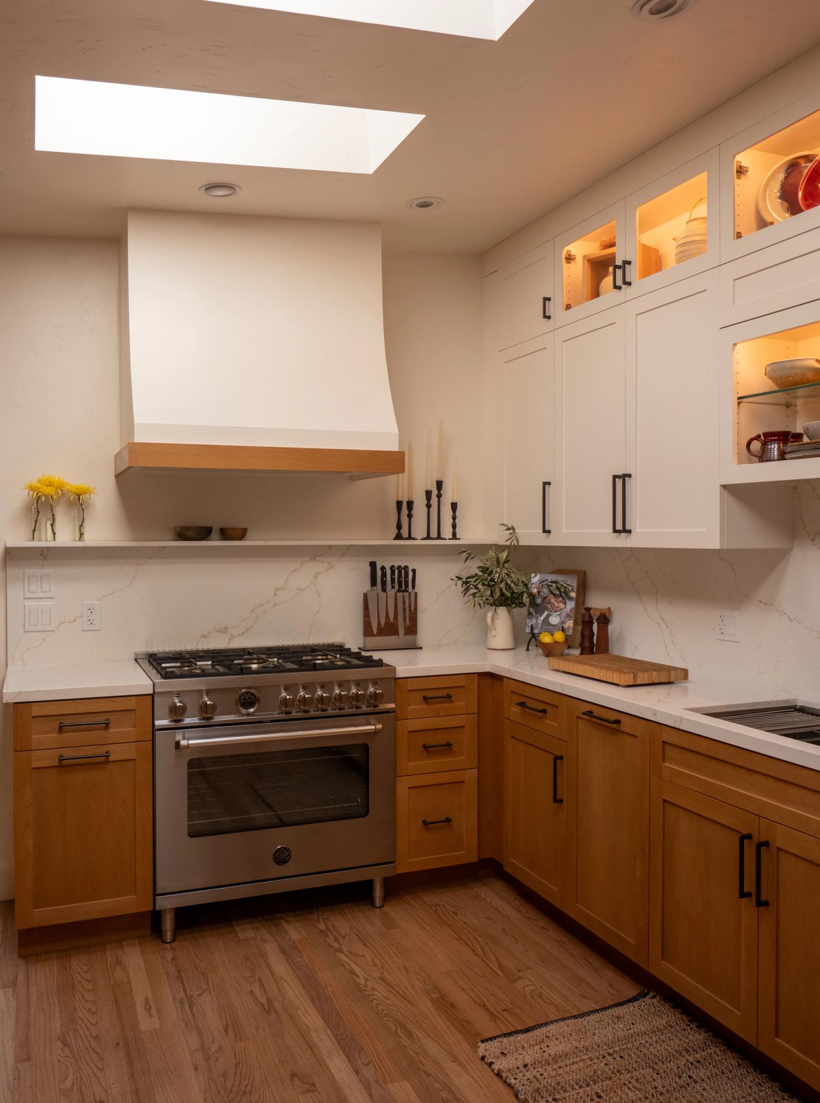 A warm transitional kitchen featuring honey-toned lower cabinets and white upper cabinets with interior lighting, a stainless steel professional gas range, marble-look quartz backsplash and countertops, and a large skylight overhead.